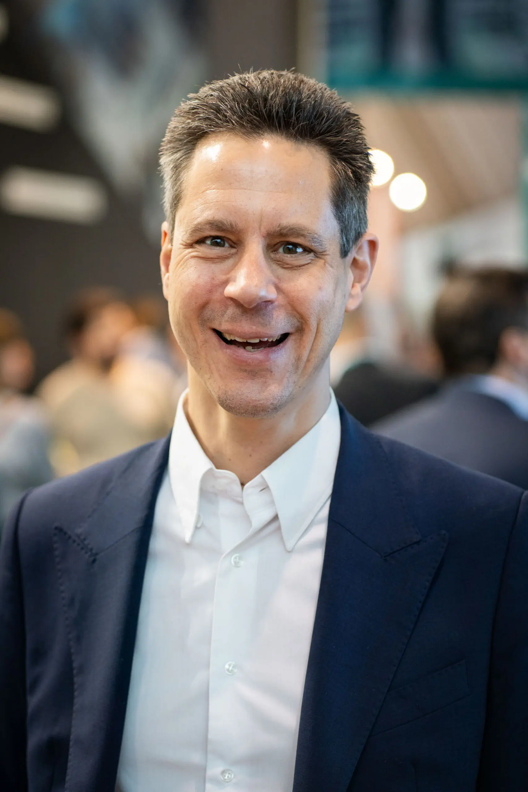 Smiling person in suit at event with blurred crowd.