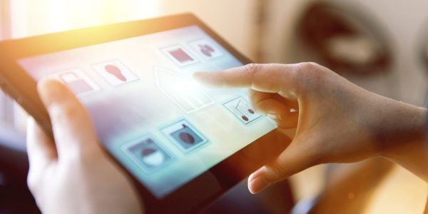 Woman puts her finger on an icon of a house shown on the touch screen of a digital tablet. The tablet runs a smart home app. Concept of controlling wireless devices at home, also know as internet of things.